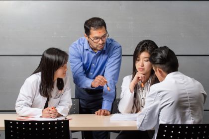 a professor stands next to a table with three seated students