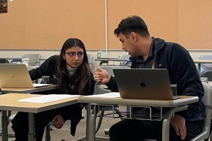 two students sit at desks while talking to each other