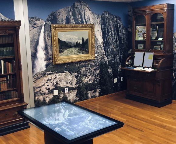 a room with an antique desk and bookcase filled with old books, a painting of the Sierra Nevadas, and a digital screen for viewing information